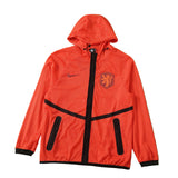 A bright orange hooded windbreaker jacket with black zippers and trim, featuring a Nike swoosh and the Netherlands national team crest, laid flat on a green soccer field background.